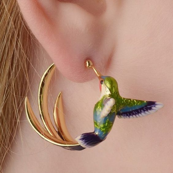 A person wearing a gold-colored hoop earring with an enamel hummingbird charm attached to it.